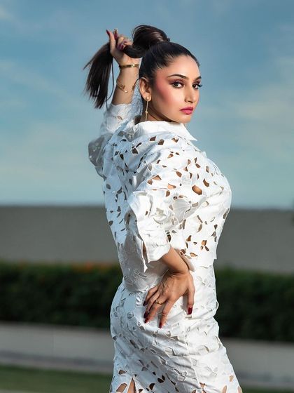 A high-fashion outdoor portrait for a magazine. The pose and high ponytail hairstyle complement the structured, modern design of the white laser-cut outfit.