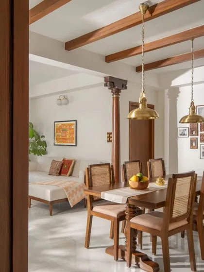 The dining area is defined by a custom teak table with cane-back chairs and accented by brass pendant lights. A traditional wooden column separates the dining space from the family living area.