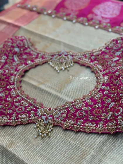 The back of a rani pink bridal blouse, featuring a heart-shaped cutout lavishly framed with intricate zardosi, stone work, and pearl tassels.
