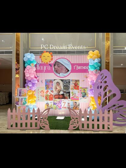 This personalized photo backdrop for Aarya's first birthday features a timeline of her first year. The setup is framed by pastel balloon columns and a large butterfly wing cutout, creating a sweet and memorable photo spot.