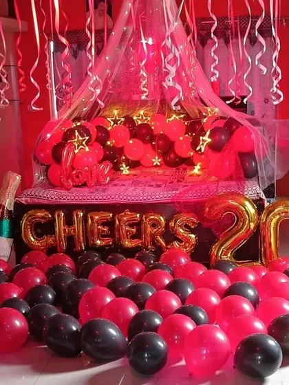 A full view of a red and black themed party decoration. The setup includes a champagne bottle prop, 'CHEERS' foil balloons, and a bed canopy with star-shaped lights, perfect for a 20th anniversary or birthday.