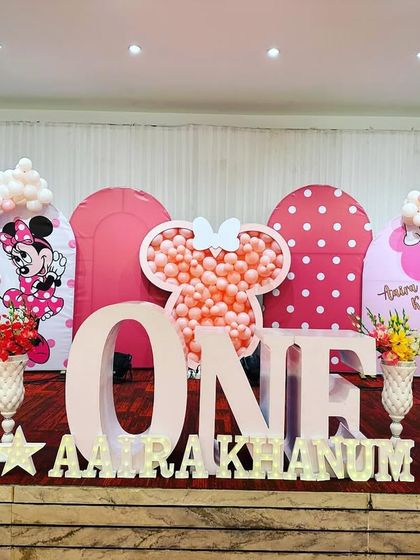 A full stage view of a Minnie Mouse first birthday party. The large 'ONE' letters are a centerpiece, surrounded by pink backdrops and a custom Minnie-head balloon frame.