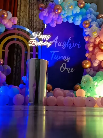 This moody and modern first birthday setup for Aashvi uses a dark backdrop to make the colors pop. The decor includes a rainbow arch cutout, a personalized neon sign, and a balloon garland in a unique palette of blue, pink, and copper.