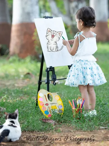 A little girl painting a picture of a cat, with a real cat as her audience.