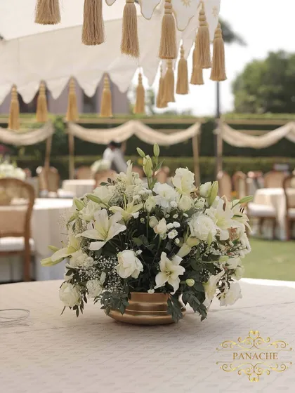 A close-up of a floral centerpiece, a lush arrangement of white lilies and roses in a golden vase, placed under a tasseled canopy.