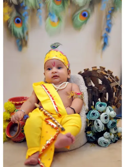 A detailed shot of a 4-month-old baby in a yellow Krishna costume, complete with a flute and peacock feather crown. The setup includes a spinning wheel prop and floral decorations.