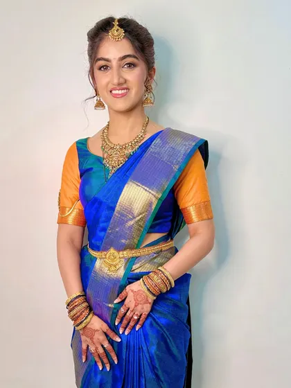 A full-length portrait showcasing a classic South Indian bridal stance. The makeup is elegant and understated, allowing the rich saree and jewellery to take center stage.