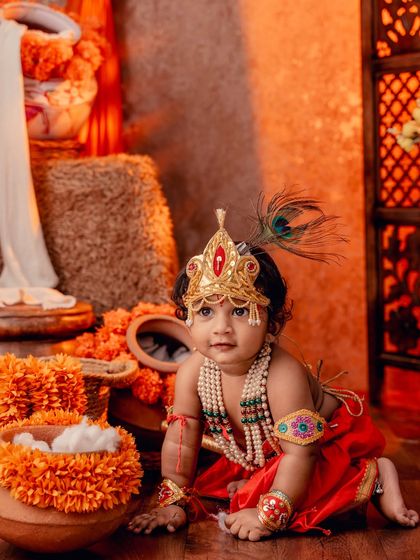 A crawling Little Krishna, looking curious and adorable in his red costume amidst a festive orange setting.