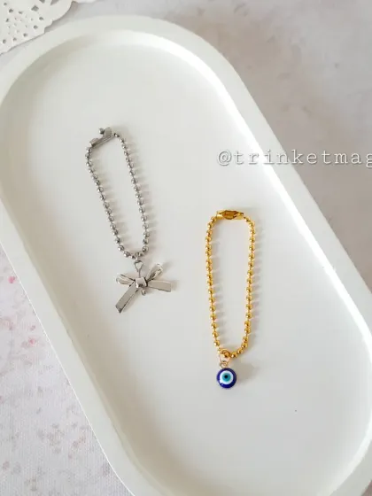 This image showcases two minimalist ball-chain charms. One features a silver bow, and the other has a gold chain with a protective evil eye, perfect for a subtle statement.