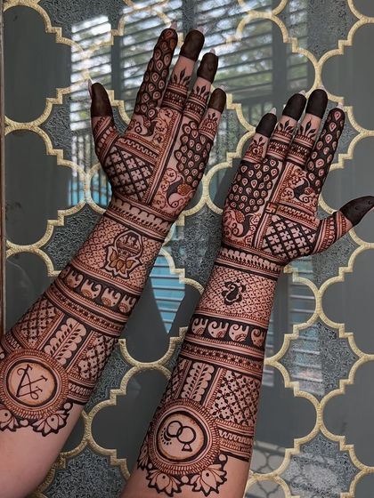 A clear, detailed shot of the engagement mehendi against a patterned background. You can clearly see the elephant procession, the couple's initials, and the ring symbols.