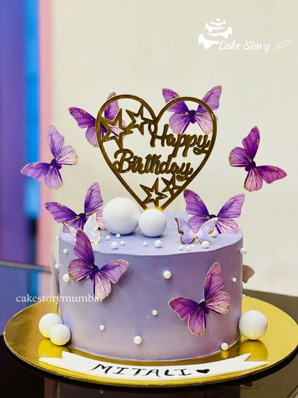 A beautiful purple butterfly-themed cake. The soft purple cake is adorned with fluttering butterflies and a heart-shaped 'Happy Birthday' topper.
