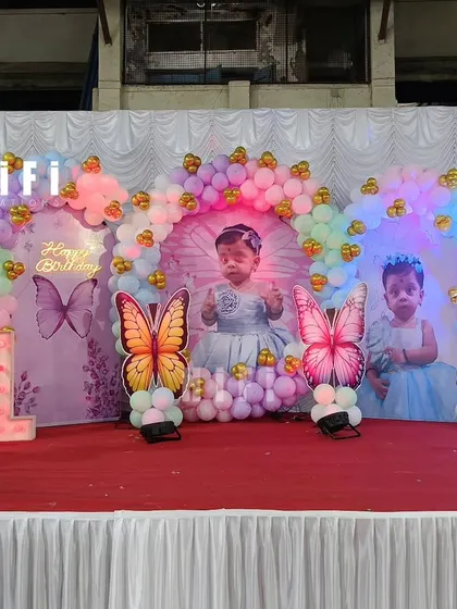 A magical butterfly-themed first birthday stage. The backdrop features photos of the birthday girl, surrounded by a pastel balloon arch and colorful butterfly cutouts.