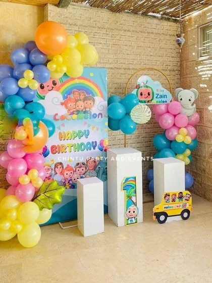A Cocomelon themed first birthday party. This setup features a colorful backdrop with characters, a balloon garland, and a '1' prop made of wood.
