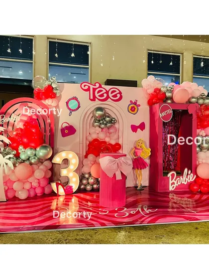 A full view of the vibrant Barbie party stage, showcasing the custom "Tee" branding and various themed icons like purses and mirrors.