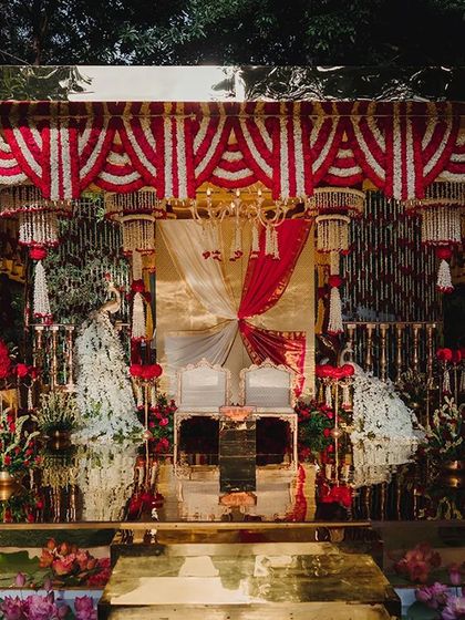 A complete view of the splendid decor, where every detail is a testament to love. The mandap features red and white floral thorans, radiant lamps, and peacocks with white floral tails for a touch of regal splendor.