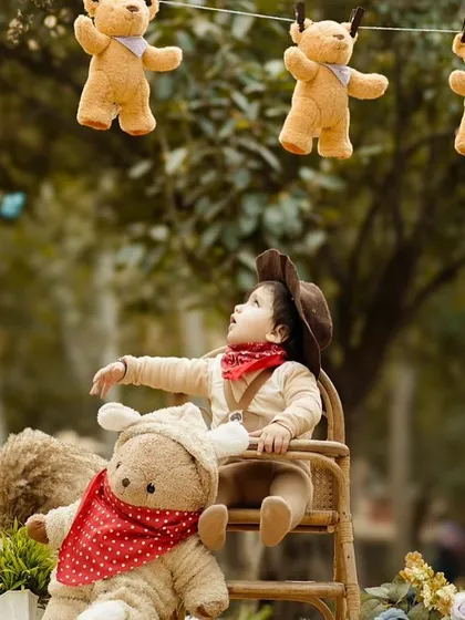 A baby dressed as a little cowboy looks up at a line of hanging teddy bears during an outdoor shoot. This creative, themed session combines the fun of props with a natural, park-like setting.