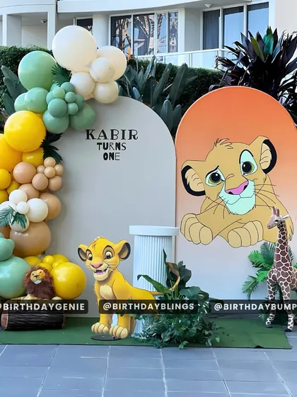 An outdoor Lion King party setup, perfect for a sunny day. This design uses two arched backdrops, one with a large Simba graphic, and a beautiful organic balloon garland in safari-inspired colors.