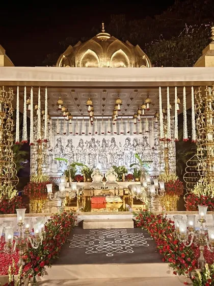 A frontal view of the magnificent brass mandap, showcasing its intricate design and symmetry. The backdrop features a hand-drawn illustration of deities, while the foreground is filled with red roses and traditional brass lamps.