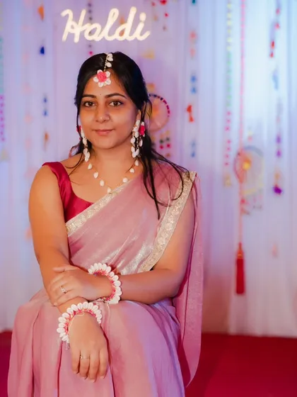 A full-length portrait of the bride at her Haldi ceremony, seated in front of the decorated backdrop. She is dressed in a lovely pink saree with floral jewelry, looking serene and beautiful.