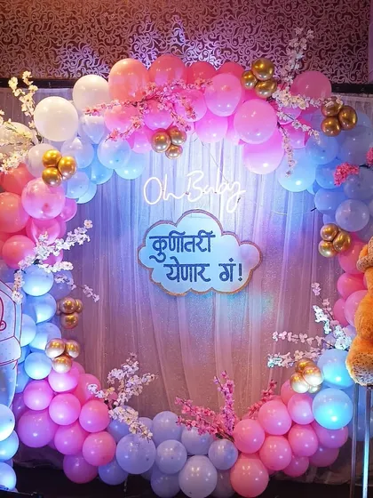 A full balloon ring decoration for a Marathi baby shower. The design incorporates the traditional 'Kunitari Yenar Ga!' sign, a teddy bear, and baby cutouts within a pink and blue balloon circle.