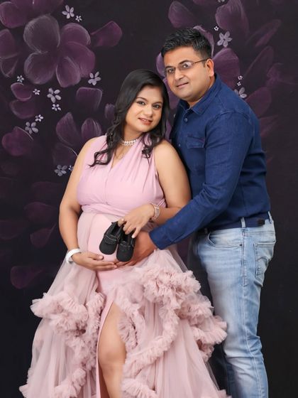 A happy couple poses with a pair of baby shoes in the studio. The ruffled pink gown and floral background create a soft and celebratory atmosphere for this pregnancy portrait.