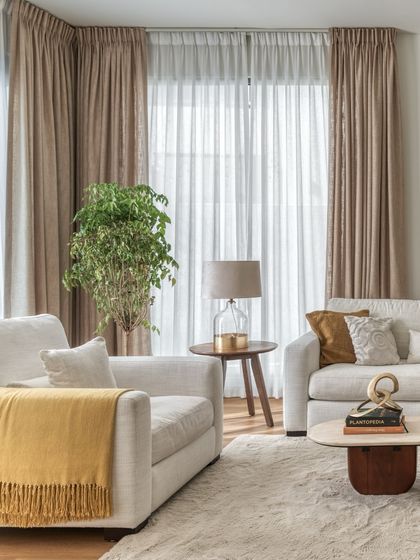 Soft, layered curtains diffuse the strong Dubai sun, filling the living room with a gentle, ambient light. Pops of yellow in the throw blanket add a touch of warmth and cheerfulness to the neutral palette.