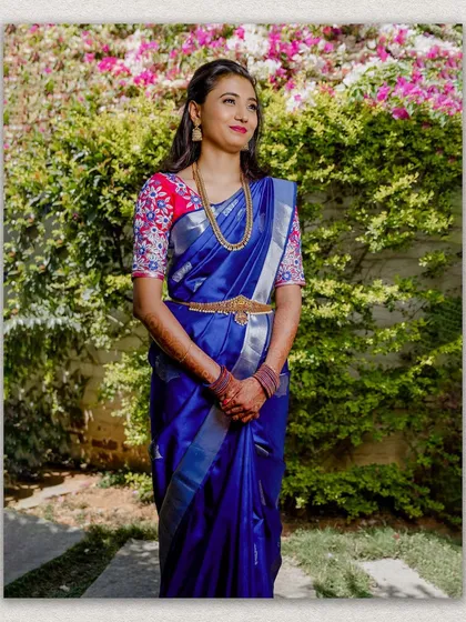 A lovely outdoor portrait of a bride in a vibrant blue and pink saree. The natural background of green foliage makes the colors of her outfit pop, capturing a fresh and happy wedding day moment.