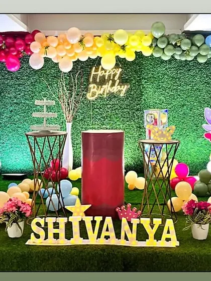 A rainbow fairy garden party with a grass wall and multi-colored balloon arch. The setup features several fairy cutouts and a neon 'Happy Birthday' sign.