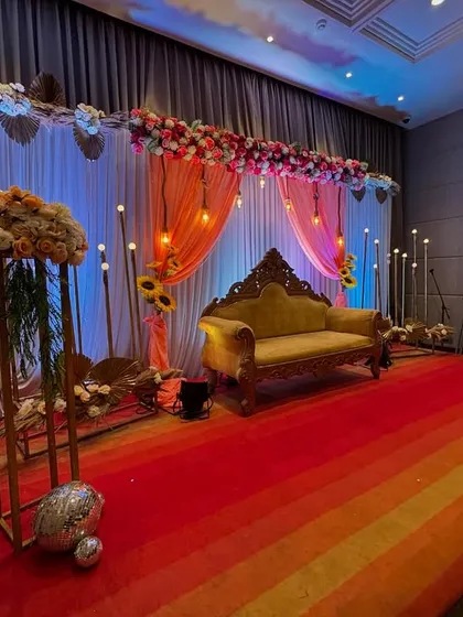 This is one of our elegant stage designs for an engagement or anniversary celebration. It features a classic carved sofa, a colorful striped carpet, and a backdrop of peach and white drapes with hanging vintage bulbs. Floral arrangements and modern light stands complete this sophisticated look.