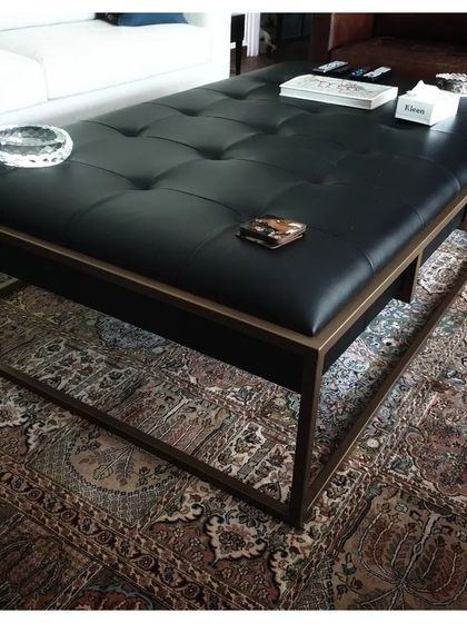 This piece is a hybrid of a coffee table and an ottoman, designed for comfort and practicality. It features a button-tufted black leather top set within a sleek metal frame, offering a versatile centerpiece for a living room.