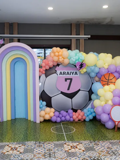 A sports-themed 7th birthday party combining a love for football with other games. The setup features a large soccer ball, a pastel rainbow arch, and cutouts of other sports equipment.