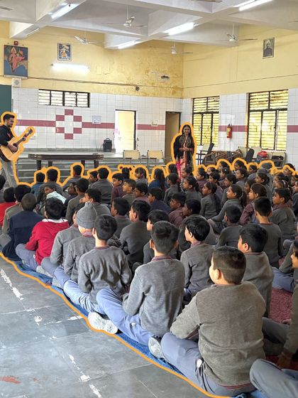 A large group of students sits attentively on the floor, engaged in a music session led by our fellows. This setup fosters a sense of community and shared experience, making every child feel part of the music.