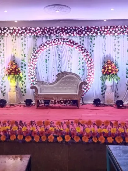 This reception stage combines a circular floral arch with a classic white chaise lounge. The backdrop of white curtains with hanging green vines adds a touch of nature, while the marigold border at the front provides a traditional element.