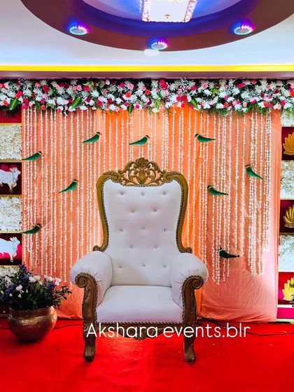 A festive Seemantham stage with a backdrop of red, yellow, and white panels. The decor includes parrot motifs and a shimmering central curtain, creating a vibrant and joyful atmosphere.