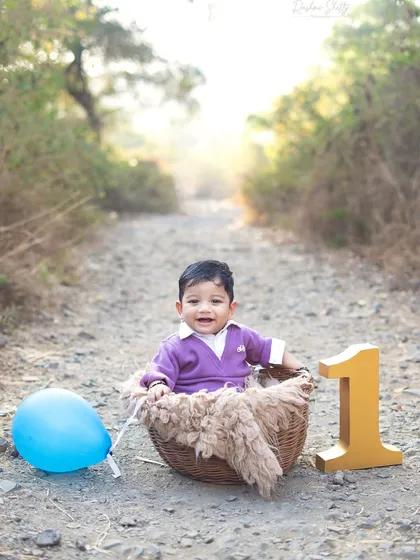 A happy one year old sitting in a basket on a rustic path. The warm, golden hour sunlight adds a magical glow to this outdoor milestone portrait.