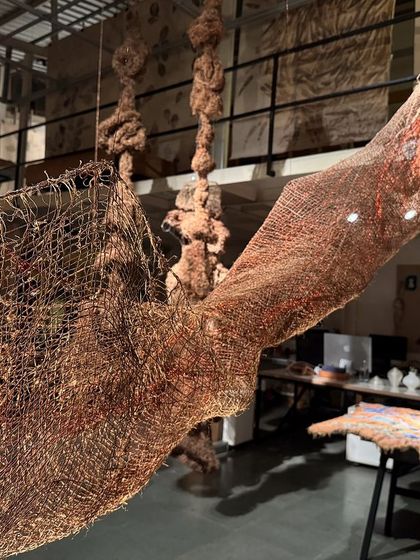A large, suspended sculpture from 'Cu+Fibers' made from woven copper and jute. The piece hangs like a chrysalis, its form shaped by the interplay of light and the inherent properties of the materials.