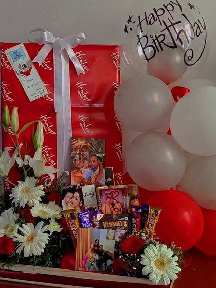 A classic red and white themed birthday hamper that combines elegance and fun. It features a large, wrapped gift from Hamleys, fresh flowers, chocolates, and personalized photos for a complete birthday surprise.