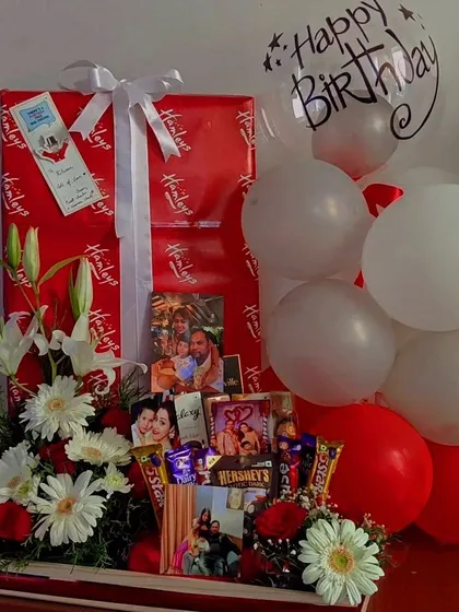A classic red and white themed birthday hamper that combines elegance and fun. It features a large, wrapped gift from Hamleys, fresh flowers, chocolates, and personalized photos for a complete birthday surprise.