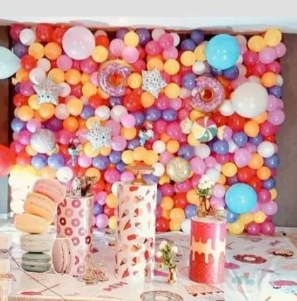 A vibrant and colorful donut-themed first birthday party. The backdrop is a dense wall of multi-colored balloons, with fun donut props and 'ONE' letters.