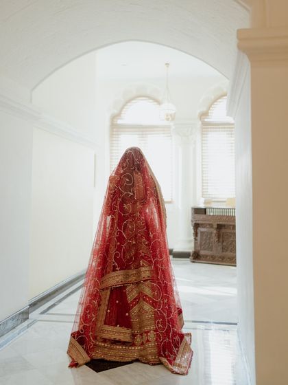 A mysterious and beautiful shot of the bride from behind, her face obscured by her red veil. Walking down a white corridor, the image is full of anticipation and traditional elegance.