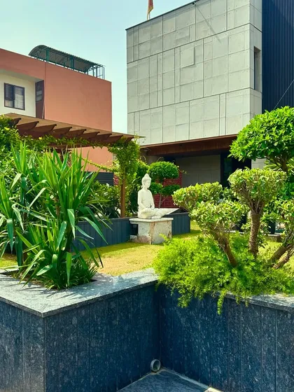 A wider view of the Faridabad garden, showing the Buddha statue as the central focal point, surrounded by lush greenery and a variety of plant forms.