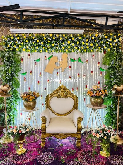 An elegant baby shower setup featuring a white backdrop with hanging jasmine and red flower buds. The decor is enhanced by parrot motifs and a central cutout symbolizing mother and child.