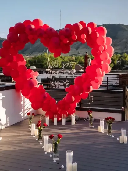 A stunning rooftop proposal setup waiting for the special moment. The focal point is a large heart-shaped arch made of red balloons framing a "Will you marry me?" neon sign against the evening sky.