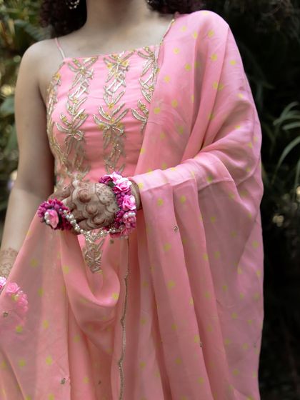 A detail shot of the bride's pink Haldi outfit and floral jewelry. The focus on her hands and the delicate floral bracelets highlights the beauty of her look.