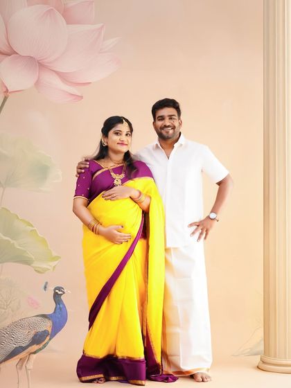 A full-length portrait of a couple during their traditional maternity shoot, set against a beautifully painted backdrop with a peacock and lotus flowers.