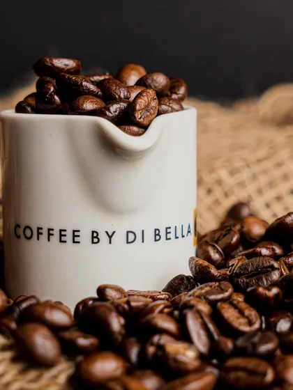 This close-up shot focuses on the rich, dark coffee beans, using a branded ceramic pitcher. The rustic burlap background adds a warm, earthy texture that speaks to the coffee's quality.