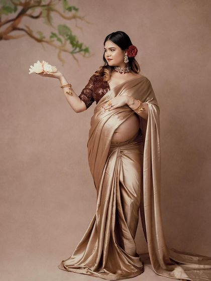 A graceful portrait of a mom-to-be in a golden saree, holding a white lotus. The simple, elegant pose and the soft lighting create a serene and beautiful image.