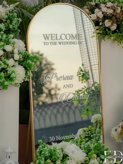 An elegant welcome sign for a botanical-themed wedding, featuring a gold-framed mirror with custom calligraphy. The sign is surrounded by lush arrangements of white and green flowers, setting a sophisticated tone.
