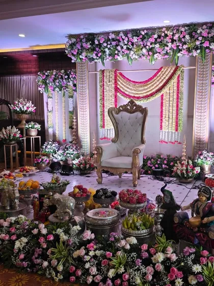 An elegant stage setup for a baby shower or engagement, featuring pillars wrapped in white flowers and a backdrop of pink and white floral garlands. The central throne and surrounding flower arrangements create a classic look.