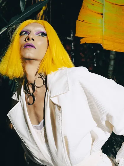 Using flash to create a stark, high-contrast image. The vibrant yellow hair and purple makeup pop against the dark, natural foliage, creating a surreal and captivating fashion portrait.